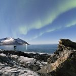 Photographer under the stars and Northern Lights (aurora borealis) surrounded by rocky peaks and icy sea, Tungeneset, Senja, Troms, Norway, Scandinavia, Europe